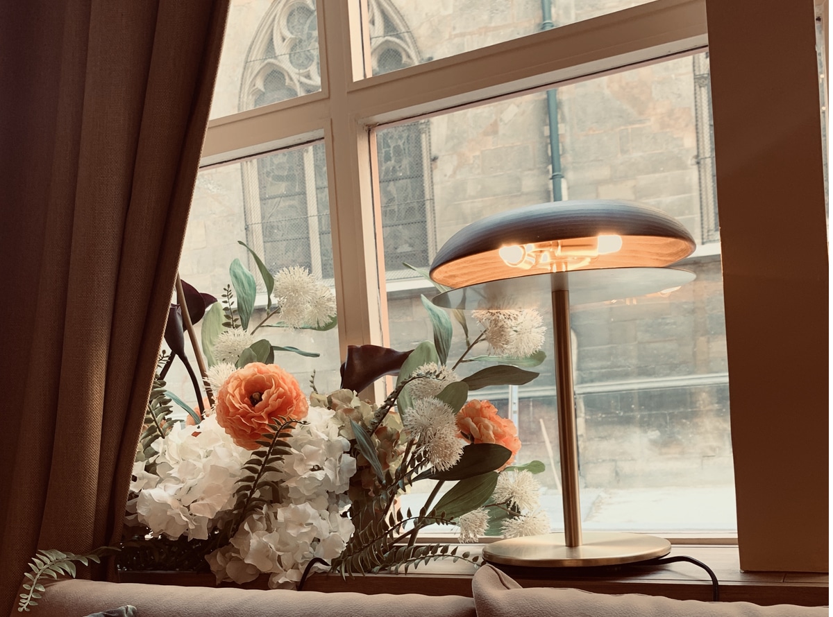 Lampe de table moderne éclairant un bouquet de renoncules et d'hortensias sur un rebord de fenêtre. Vue sur une façade en pierre et des détails gothiques.