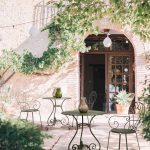 Thumbnail of http://Un%20patio%20extérieur%20rustique%20avec%20des%20tables%20et%20des%20chaises%20en%20fer%20forgé,%20chaque%20table%20étant%20ornée%20d'un%20petit%20vase.%20Le%20patio%20est%20ombragé%20par%20un%20grand%20arbre%20et%20encadré%20par%20un%20bâtiment%20en%20pierre%20avec%20des%20portes%20cintrées%20et%20du%20lierre.%20Des%20lumières%20suspendues%20ajoutent%20une%20touche%20de%20charme.