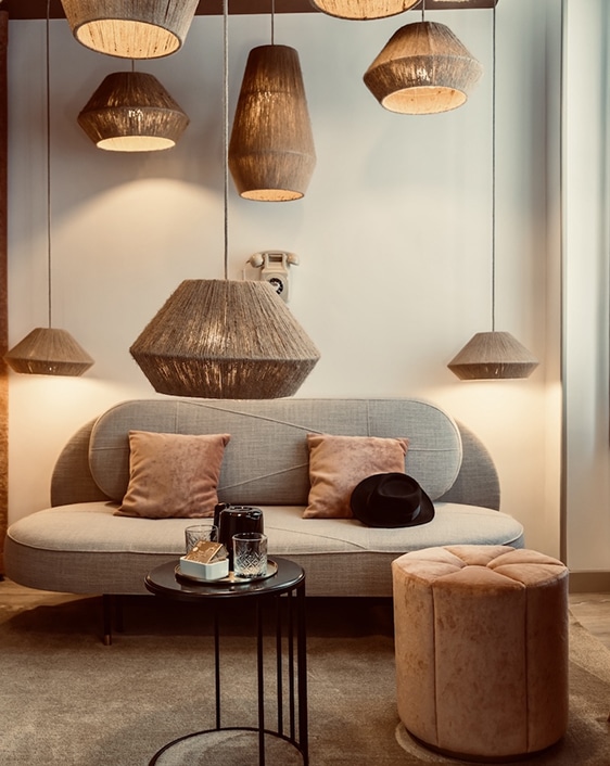 Suspension fibres naturelles : ambiance cosy dans un salon design. Salon design avec canapé courbé, pouf rose et table basse. Des suspensions en fibres naturelles de formes variées éclairent un téléphone vintage mural.