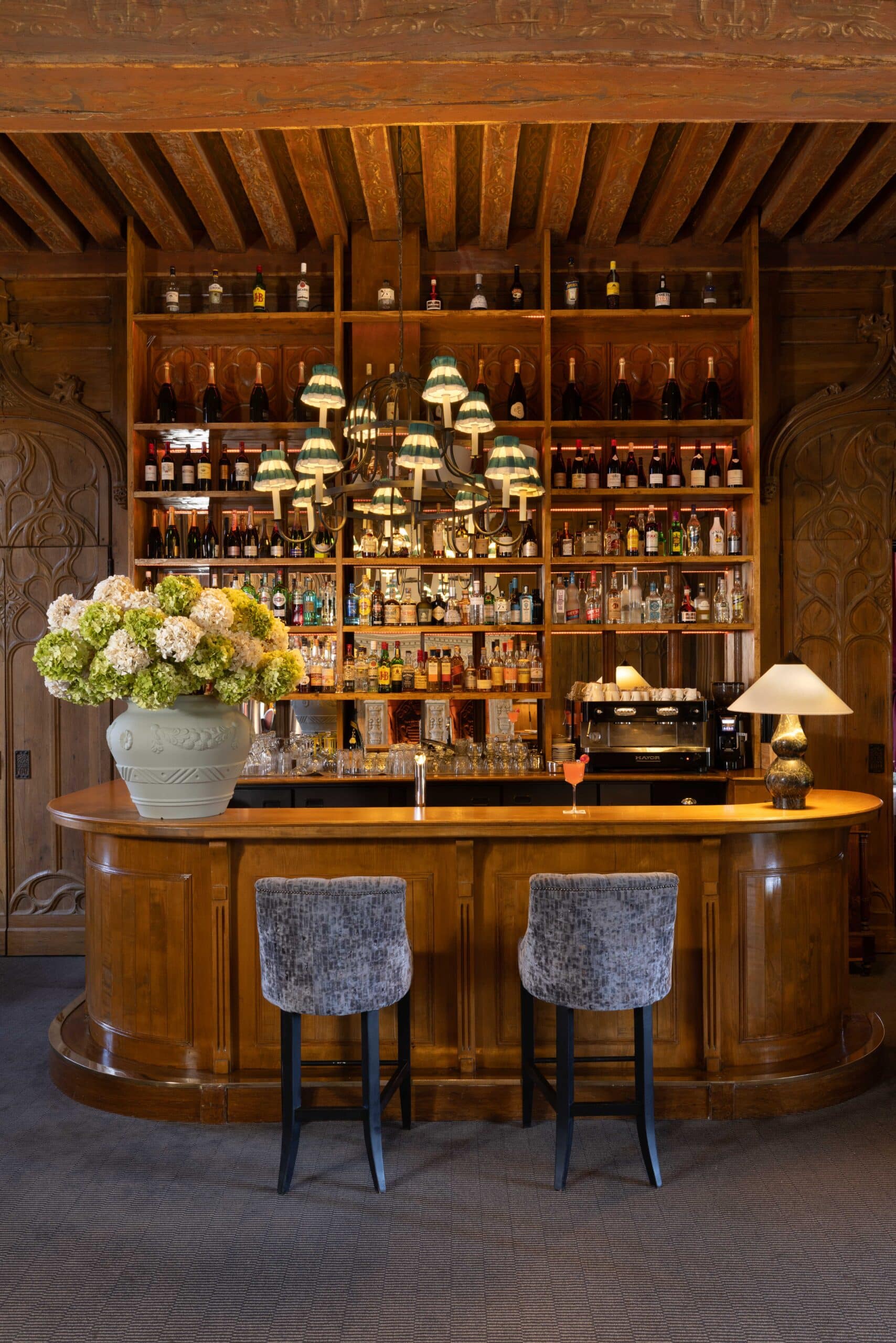 Luxueux bar de château en bois sculpté, plafond à poutres peintes. Grand chandelier à abat-jour rayés, étagères d'alcool et tabourets.