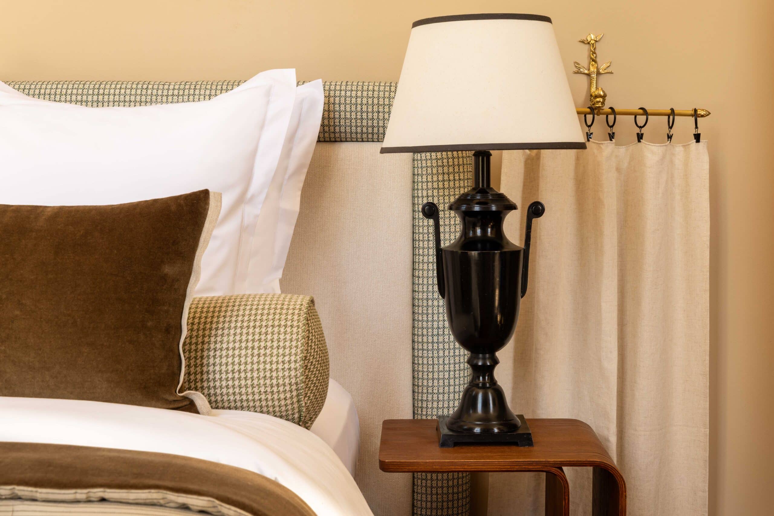 Chambre élégante: tête de lit texturée, coussins marron et pied-de-poule. Lampe noire urne et tringle à rideau dorée ornementée.