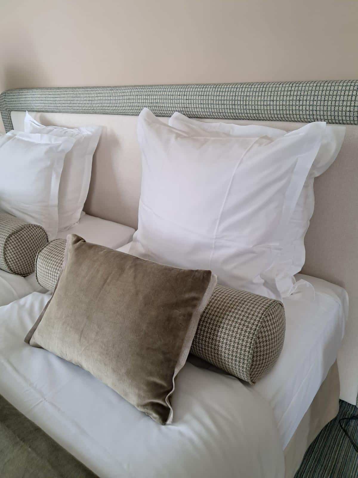 Lit élégant avec tête de lit beige et traversin à motif houndstooth vert-gris. Draps blancs et coussin central en velours taupe.