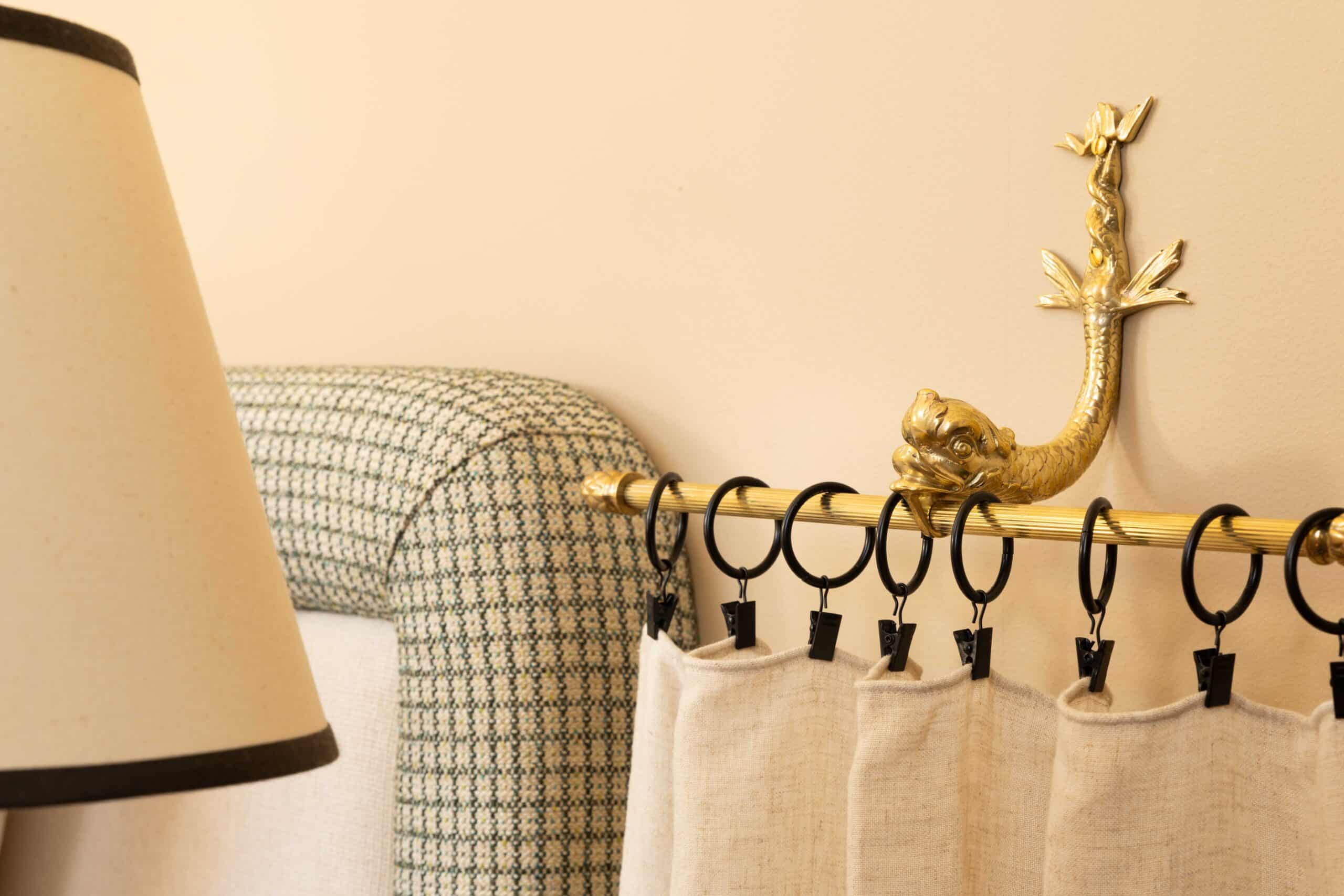 Détail de tringle à rideau en laiton avec finial dauphin, rideaux en lin brut et tête de lit texturée.