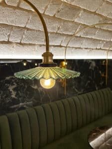 Suspension en verre plissé vert avec bras doré devant un mur en marbre noir et une banquette en velours vert.